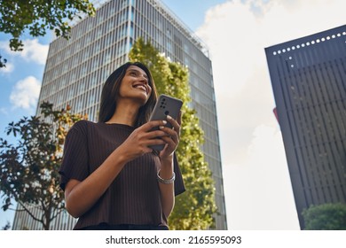 Smiling beautiful businesswoman standing against office building with mobile phone - Shutterstock ID 2165595093