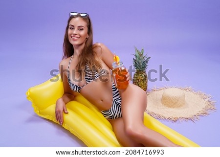 Smiling beautiful caucasian adult girl wears in bikini lyes on inflatable yellow matress while holds cocktail on purple background . Summer concept. Travel concept. Relax concept 