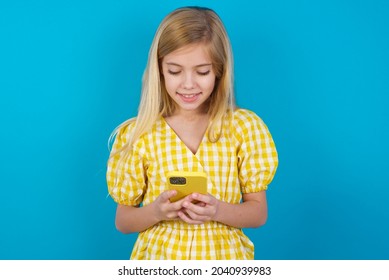 Smiling Beautiful Caucasian Little Girl Wearing Yellow Dress Over Blue Background Using Cell Phone, Messaging, Being Happy To Text With Friends, Looking At Smartphone. Modern Technologies 