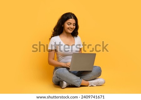 Smiling beautiful curly young indian woman wearing casual clothing enjoying newest educational course, typing on laptop computer, sitting on floor over yellow studio background
