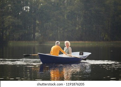 Smiling beautiful curly-haired senior lady in cardigan sitting on boat and talking to boyfriend while he rowing oars - Powered by Shutterstock