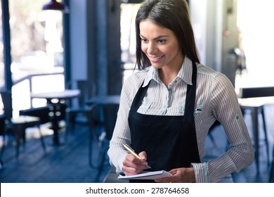 Smiling beautiful female waiter in apron with notepad and pen in cafe. Looking at camera - Shutterstock ID 278764658