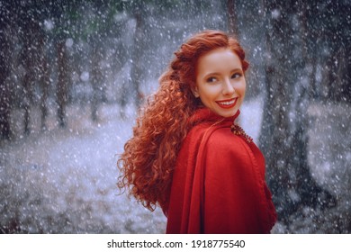 Smiling Beautiful Girl With Red Curly Hair In Red Cloak Walk In Winter Forest. Historical Reconstruction Of Ancient Celtic Times.