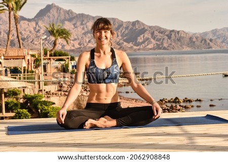 Smiling beautiful girl sittin in yoga pose on a sandy beach with rocky mountains on background on sunny summer day enjoying momets of sunrise