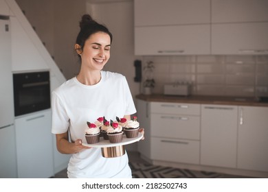 Smiling beautiful happy woman making tasty homemade cupcakes with flower rose decor holding tray with pastry at home kitchen.  - Powered by Shutterstock