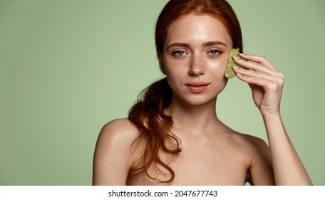 Smiling beautiful head and shoulders of redhead girl with freckles using facial gua sha jade board isolated on green background