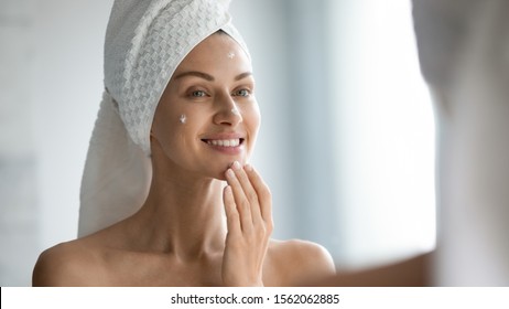 Smiling beautiful lady apply skincare cream on face look in bathroom mirror, happy young woman wrap towel on head put facial creme on doing morning healthy skin care beauty routine treatment concept