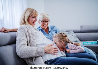 Smiling beautiful pregnant woman holding belly and sitting on sofa in living room while her daughter kissing belly. Next to pregnant woman is her cheerful proud mother.