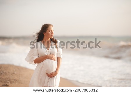 Smiling beautiful pregnant woman wear stylish white dress hold belly walk at beach over waves of sea outdoors. Looking away. Motherhood. Maternity. Healthy lifestyle. Happiness.