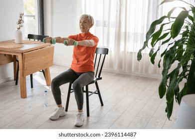 Smiling beautiful senior woman health instructor doing chair exercises with dumbbells - Powered by Shutterstock