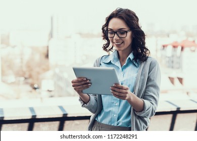 Smiling Beautiful Woman Holding Tablet Outdoor. Portrait Of Beautiful Business Woman Wearing Glasses Standing Outside Building And Using Tablet. Happy Attractive Woman Reading From Tablet