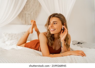 Smiling Beautiful  Woman Listening Lovely Music  While Lying On Stomach  In Bed. Warm Colors. Home Cozy Atmosphere. 