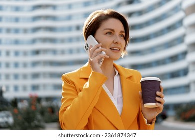 Smiling  beautiful  woman with short haircut  in yellow jacket and pants using mobyle phone , holding cup of coffe . Standing over modern city.  - Powered by Shutterstock