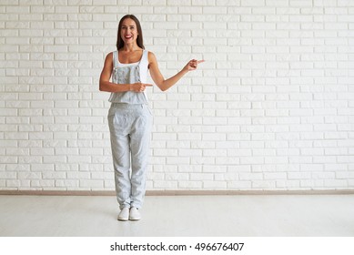 Smiling beautiful woman standing and points with both hands to the side, white brick wall on background