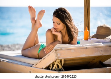 Smiling beautiful woman sunbathing in a bikini on a beach at tropical travel resort,enjoying summer holidays.Young woman lying on sun lounger near the sea.Happy serene woman having SPF protection