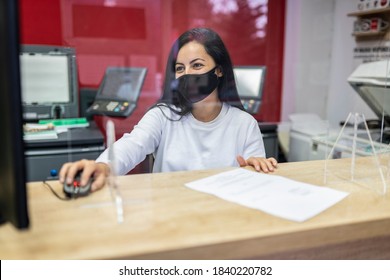 Smiling beautiful woman wearing face mask and using computer in copy shop, plastic transparent barrier between her and customer