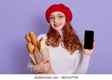 Smiling beautiful woman wearing white shirt and red beret, looking at camera with happy facial expression, ginger girl with baguettes in hands, female showing blank screen of smart phone.