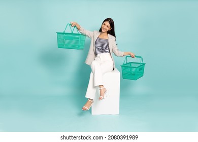 Smiling Beautiful Young Asian Businesswoman In Suit Holding Shopping Basket And Sitting On White Box Isolated On Green Background