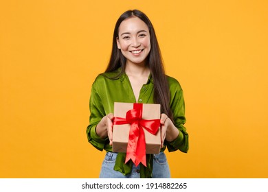 Smiling beautiful young brunette asian woman wearing basic green shirt standing hold red present box with gift ribbon bow looking camera isolated on bright yellow colour background, studio portrait
