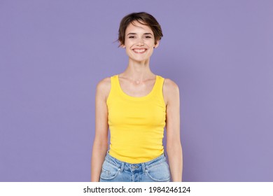 Smiling beautiful young brunette woman girl wearing yellow casual tank top posing isolated on pastel violet wall background studio portrait. People sincere emotions lifestyle concept. Looking camera