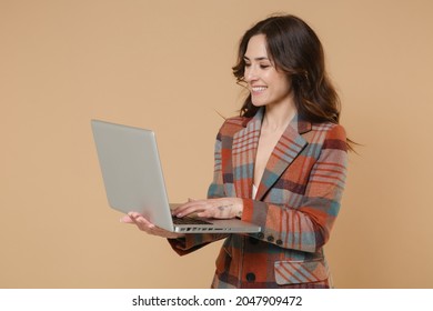 Smiling beautiful young brunette woman 20s wearing casual checkered jacket posing standing holding in hands working on laptop pc computer isolated on pastel beige colour background, studio portrait