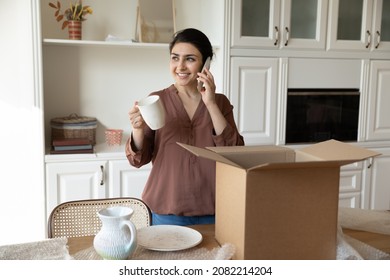 Smiling Beautiful Young Indian Ethnic Woman Homeowner Holding Phone Call Conversation, Sharing Positive Shopping Experience, Unpacking Dinnerware Set From Huge Cardboard Box In Modern Kitchen.