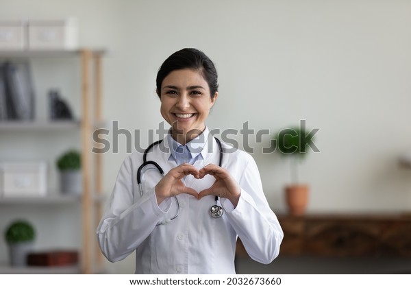 Smiling\
beautiful young indian ethnicity female doctor cardiologist showing\
heart symbol with fingers, expressing love and support to patients,\
healthcare medical help charity donation\
concept.