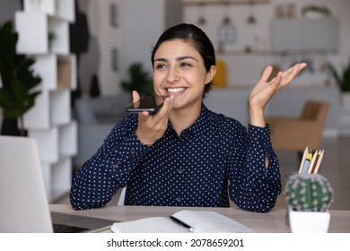 Smiling beautiful young Indian woman dictating audio message, holding loudspeaker conversation, web surfing information online, using ai assistant app or voicemail on cellphone at home office.