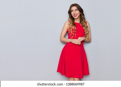 Smiling beautiful young woman in elegant red dress is posing and looking away. Three quarter length studio shot on gray background.