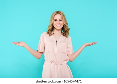 Smiling Beautiful Young Woman In Pale Pink Smart Dress Presenting Product And Looking At Camera. Three Quarter Length Studio Shot On Pastel Blue Background.