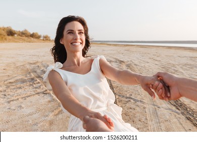 Smiling beautiful young woman wearing summer dress holding her boyfriends outstretched hand while walking at the beach