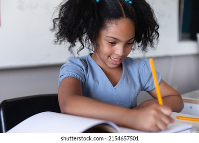 Smiling biracial elementary schoolgirl writing on book while studying at desk in classroom. unaltered, education, learning, studying, concentration and school concept. - Powered by Shutterstock