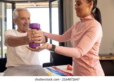 Smiling biracial female physiotherapist helping caucasian senior man in lifting dumbbell at home. Assistance, unaltered, physical therapy, exercise, healthcare, retirement, treatment, recovery. - Powered by Shutterstock