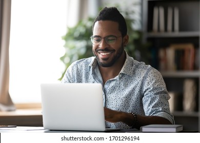 Smiling biracial man in glasses sit at desk in office browsing wireless Internet on laptop device, happy African American male worker laugh watch funny video on computer gadget, relax at work break