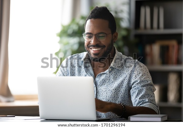Smiling biracial man in glasses sit at desk in\
office browsing wireless Internet on laptop device, happy African\
American male worker laugh watch funny video on computer gadget,\
relax at work break