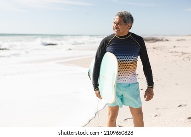 Smiling biracial senior man holding surfboard looking away at beach against sky on sunny day. water sport and active lifestyle. - Powered by Shutterstock