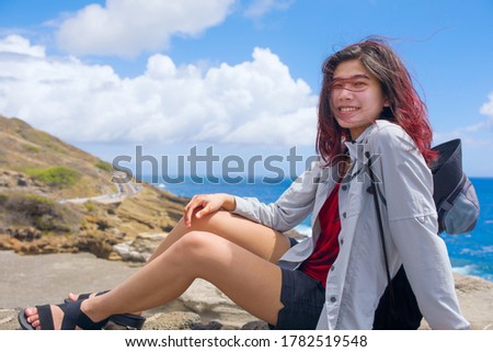 Smiling biracial teen girl sitting on edge of ricky cliff overlooking tropical blue Pacific ocean on Hawaiian island