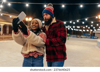 Smiling biracial young couple taking selfie via cellphone while ice skating at outdoors rink - Powered by Shutterstock