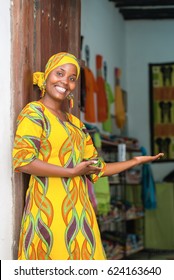 Smiling black African woman in traditional clothes wishing welcome to customers in front of her store