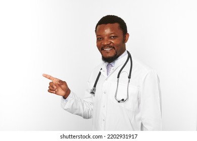 Smiling Black Bearded Doctor Man In White Coat With Stethoscope Points Finger To Left, Isolated On White Background. Adult Black African American Physician Therapist Portrait, Candid Male Emotion