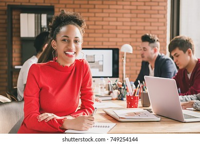 Smiling Black Business Woman In Red Top Holding Pen At Interior Design Meeting With Office Colleagues In Casual Clothes Against Brick Wall Background