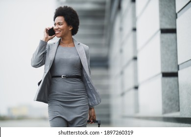 A Smiling Black Business Woman Talking On Her Smart Phone While Walking Down A Street, In Front A Corporate Building.