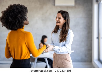 A smiling Black businesswoman and her Caucasian colleague exchange greetings with a handshake in a well-lit, contemporary workspace, symbolizing professionalism and cooperation. - Powered by Shutterstock