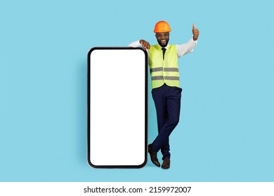 Smiling Black Construction Engineer Standing Near Big Blank Smartphone And Showing Thumb Up, African American Builder Wearing Safety Vest And Hard Hat Recommending New Mobile App, Mockup