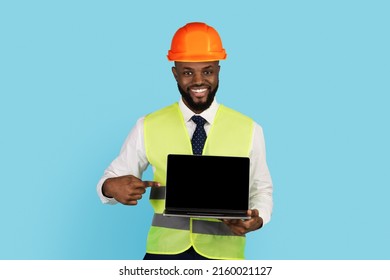 Smiling Black Construction Worker Pointing At Laptop With Blank Screen In Hand, Handsome Young African American Builder Wearing Hard Hat And Safety Vest Demonstrating Copy Space For Website, Mockup