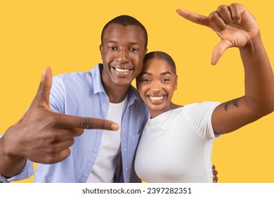 Smiling black couple playfully making camera frame with their hands, capturing moment of joy, in casual clothing against cheerful yellow background