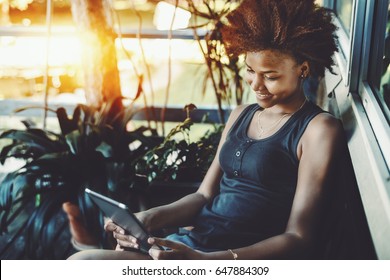 Smiling Black Curly Teen Sitting On The Porch Of Resort Summer House Near Pond And Reading Electronic Book, Cheerful Afro American Girl In T-shirt Is Making Selfie On Digital Tablet Near House Window