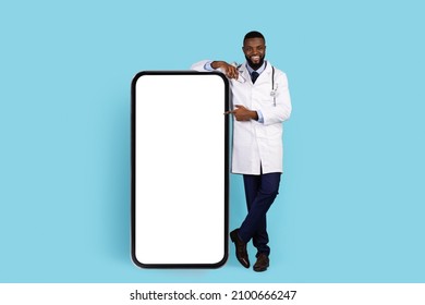 Smiling Black Doctor In Uniform Pointing At Big Blank Phone With White Screen, African American Therapist Demonstrating Copy Space For Online Medical App Design, Blue Studio Background, Mockup
