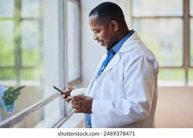 Smiling Black Doctor Using Smartphone While Holding Coffee Cup in Hospital Corridor - Powered by Shutterstock