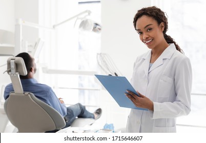 Smiling Black Female Dentist Filling Medical Form In Cabinet With Male Patient, Free Space